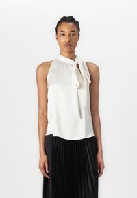 Haut en satin blanc à col halter avec un détail à nouer à l'avant, tombant doucement sur le torse. Porté sur une jupe plissée noire.