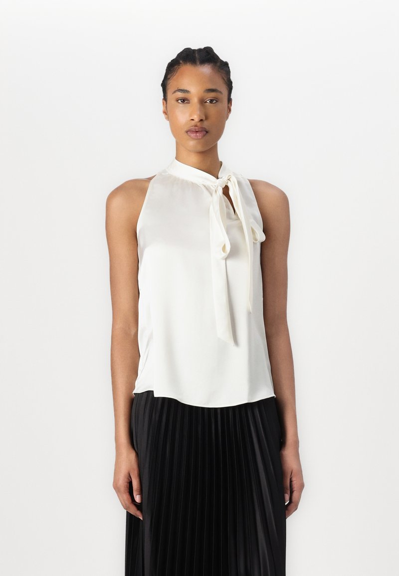 Haut en satin blanc à col halter avec un détail à nouer à l'avant, tombant doucement sur le torse. Porté sur une jupe plissée noire.
