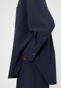 Camicia blu navy a maniche lunghe in tessuto strutturato, con una tasca sul petto e polsini con bottoni. Vestibilità morbida con spacchi laterali.