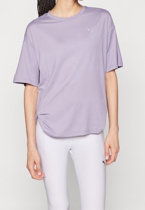 Vrouw draagt een loszittend lichtpaars t-shirt en strakke witte legging met zwart Puma-logo op linker bovenbeen, staand tegen een witte achtergrond.