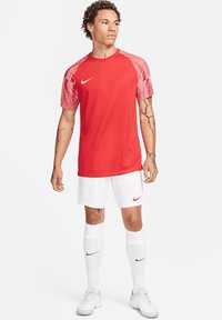 Rotes Nike Sportshirt mit gestreiften roten Ärmeln, kombiniert mit weißen Shorts und Socken. Das Model trägt weiße Sneakers.