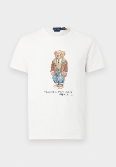 Camisola branca de algodão com uma ilustração de um urso vestido com um casaco castanho e óculos, calças de ganga e sapatos castanhos; texto "Polo Bear by Ralph Lauren".