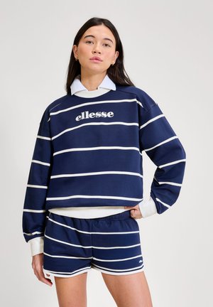 Mlada ženska, oblečena v temno modro-belo črtasto ellesse majico z dolgimi rokavi in ustrezne kratke hlače, ki stoji z roko na boku, na nevtralnem ozadju.