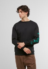 Felpa nera in cotone con collo rotondo e maniche lunghe. Presenta grafiche di palme verdi sul braccio sinistro.
