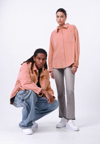 Twee modellen tonen casual outfits: een roze corduroy shirt, grijze jeans en witte sneakers. De jeans hebben een patroon in het blauw.