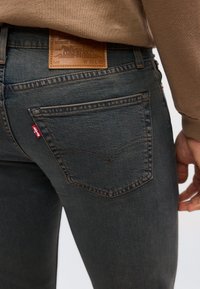 Κοντινό πλάνο της πίσω τσέπης και της μέσης μπλε τζιν παντελονιού με δερμάτινη ετικέτα μάρκας και κόκκινη υφασμάτινη ετικέτα, που φοριέται με μπεζ τοπ.