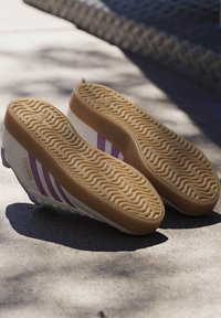 Zapatillas blancas con tres franjas moradas, suela de goma con textura en forma de ola, material superior liso, diseño plano y mínima marca.