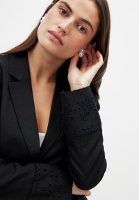 Black blazer with eyelet embroidery detail on sleeves, featuring a tailored fit and soft texture, layered over a white top.
