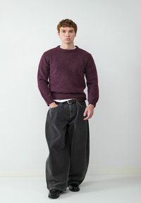 Dunkellila Strickpullover mit Rundhalsausschnitt, kombiniert mit übergroßen schwarzen Jeans und schwarzen Schuhen, vor einem neutralen Hintergrund.