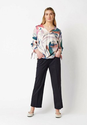 Woman standing with hands in pockets wearing a colorful abstract print blouse, black straight-leg pants, and cream flats against white background.