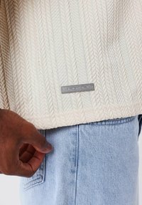 Maglione crema a maglia cable con un motivo testurizzato, caratterizzato da un etichetta grigia rettangolare sull'orlo, abbinato a jeans in denim azzurri.
