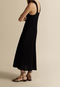 Robe midi noire à côtes avec des bretelles larges et une coupe décontractée, associée à des sandales plates noires à lanières, présentée de profil.