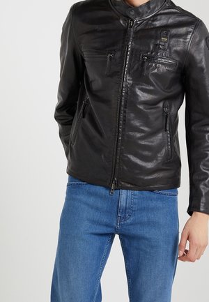 Homme portant une veste en cuir noire zippée avec plusieurs poches zippées et un jean bleu denim, debout devant un fond clair uni.