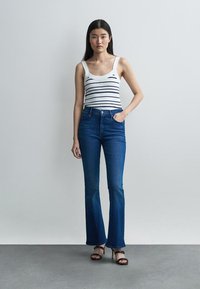 Jeune femme debout, une main dans la poche, portant un débardeur blanc à rayures bleu marine, un jean évasé taille haute bleu et des talons noirs.