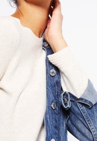 Eine Person trägt einen cremefarbenen Strickpullover und hält den Kragen einer ungerbten blauen Jeansjacke mit silbernen Knöpfen vor einem weißen Hintergrund.