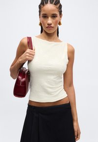 Νεαρή γυναίκα με πλεξούδες φορά αμάνικο λευκό τοπ και μαύρη φούστα, κρατώντας σκούρο κόκκινο τσάντα ώμου.