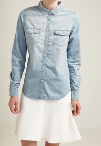 Lys blå denimskjorte med lange ærmer, knaplukning foran, spidse kraver og to brystlommer. Båret med en hvid, flagrende nederdel.