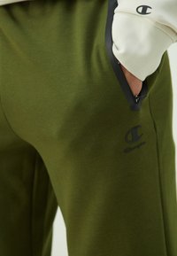 Olivgrüne Sweatpants mit glatter Textur, die ein schwarzes Logo an den Oberschenkeln und kontrastierende schwarze Akzente an den Taschenrändern aufweisen.