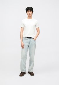 Wit T-shirt met korte mouwen, lichtblauwe jeans met rechte pijpen, bruine schoenen, staande houding, handen in de zakken, neutrale achtergrond.