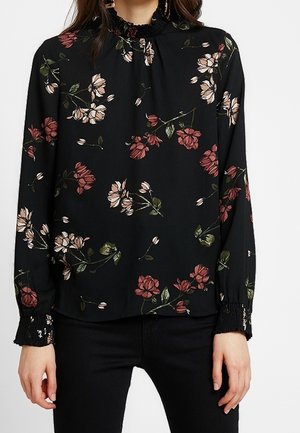 Blouse noire à manches longues avec motif floral rose et blanc, col montant à volants et poignets smockés portée avec un pantalon noir.