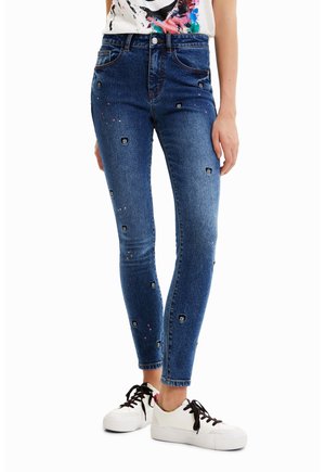 Vaqueros ajustados azules con pequeños parches negros de calaveras y salpicaduras de pintura, combinados con zapatillas blancas y una camiseta abstracta y colorida.