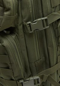 Olive green tactical backpack with multiple zippers, adjustable straps, and plastic hardware. Features a textured fabric and grid design.