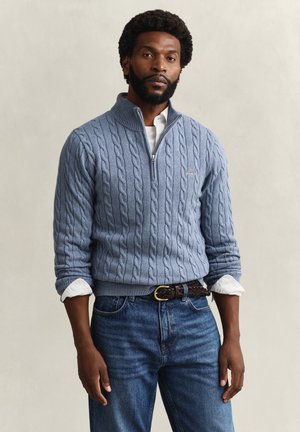 Mann med skjegg iført lyseblå kabelstrikket genser med halvlukking over hvit skjorte og blå jeans, stående foran ensfarget bakgrunn.