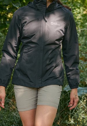 Person iført en sort Jack Wolfskin jakke med hætte og beige shorts, der går udendørs i et grønt, frodigt miljø.