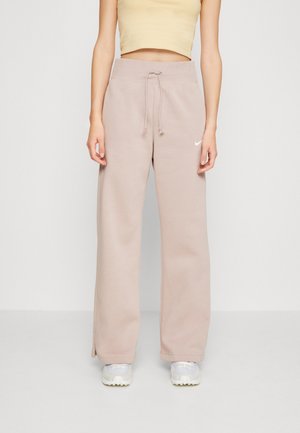 Kobieta nosząca beżowe szerokie spodnie dresowe z sznurkiem w talii i białe trampki, połączone z żółtym krótkim topem.