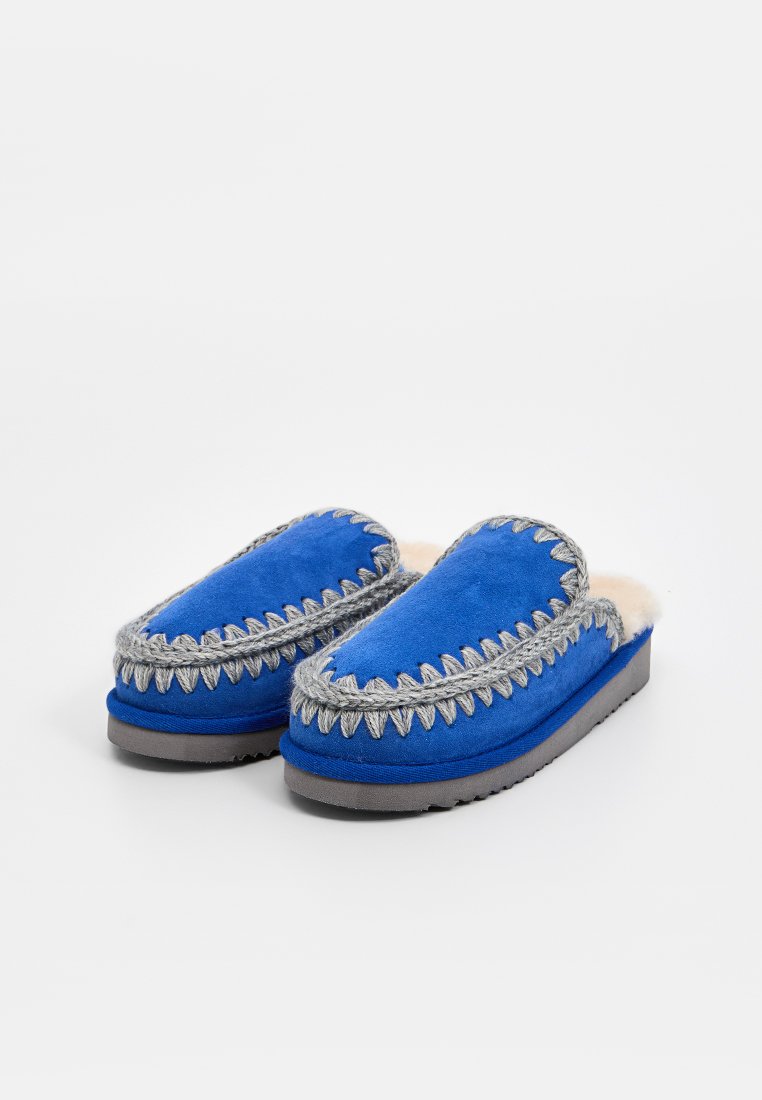 Pantofole blu senza lacci con cuciture zigzag grigie e morbida fodera interna bianca su suole grigie, posizionate su uno sfondo bianco semplice.