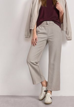 Vrouw draagt beige cropped wide-leg broek met riem, bordeaux top, lichte jas over de schouders gedrapeerd en witte sneakers met donkere strepen.