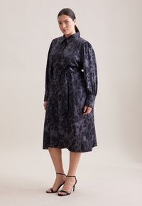 Abito camicia a fantasia scura con maniche lunghe, dettaglio in vita con laccio e texture liscia. Indossato con tacchi neri, posato su uno sfondo chiaro.