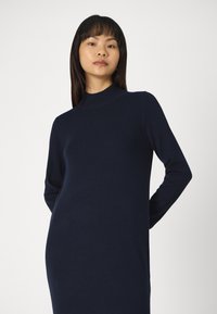 Abito a maglia blu navy con maniche lunghe e collo alto. Texture liscia e silhouette aderente, che arriva fino a metà polpaccio. Design semplice e minimale.