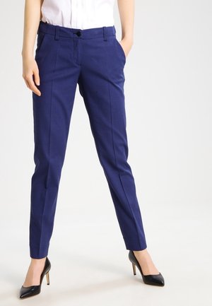 Un pantalon tailleur bleu marine avec un motif texturé, coupe slim, plis repassés et poches latérales, associé à des chaussures à talons noirs.