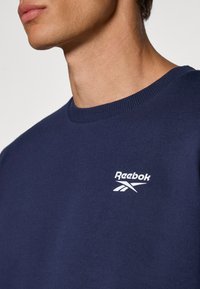 Granatowy sweter Reebok z prążkowanym dekoltem i białym logo na piersi. Materiał wydaje się miękki i strukturalny.