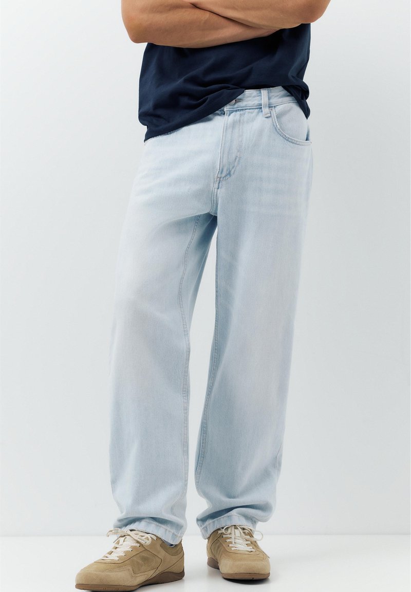 Personne portant un jean bleu clair ample, une chemise bleu marine et des baskets beiges, debout, les bras croisés, devant un fond uni.