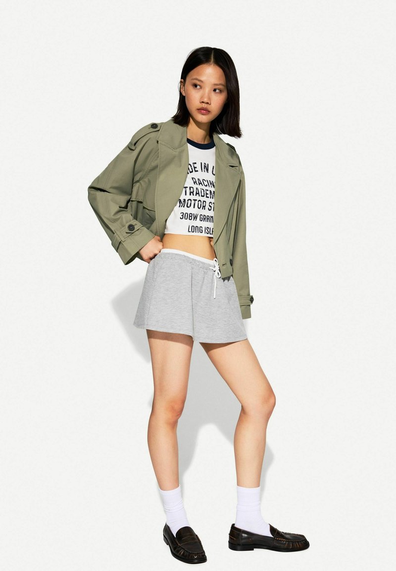 Olijfgroene trenchcoat over een witte, gekorte graphic t-shirt, gecombineerd met grijze shorts en zwarte instappers. Witte enkelkousen maken de outfit compleet.