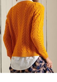 Strickpullover in leuchtendem Orange, mit einem Zopfmuster, geripptem Saum und überschnittenen Schultern, über einem weißen Hemd und einem karierten Rock getragen.