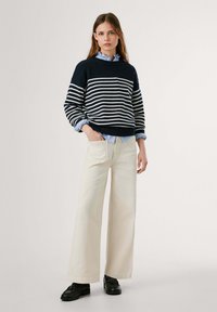 Maglione a righe navy e bianche, vestibilità ampia, abbinato a pantaloni beige larghi e scarpe nere con lacci. Camicia azzurra con colletto sotto.