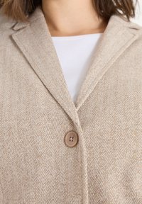 Beige blazer met een visgraatpatroon, voorzien van een geknipt revers en een enkele knoopsluiting. Zachte, gestructureerde stof met een subtiele glans.