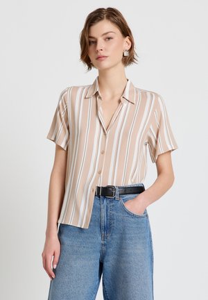 Young woman with short hair wearing beige and white vertical striped short-sleeve shirt tucked into high-waisted blue jeans with black belt.