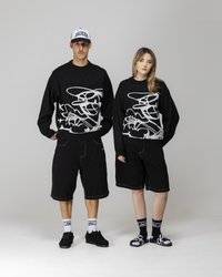 Svart oversized hettegenser med hvitt abstrakt design, kombinert med svarte shorts. Begge har på seg svarte joggesko og merkede sokker. Den ene har en hvit caps.