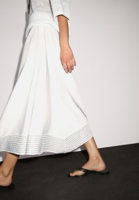 Weiße, luftige Bluse mit horizontaler gestreifter Säume, aus leichtem Material. Kombiniert mit schwarzen Flip-Flops. Minimalistisches Design vor einem schlichten Hintergrund.