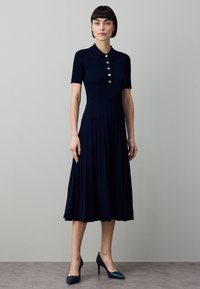 Temno modra obleka z rebrastim vzorcem, kratkimi rokavi, ovratnikom in gumbi. Razširjen, nabran krilo. Ujemajoči se čevlji z visokimi petami dopolnjujejo videz.