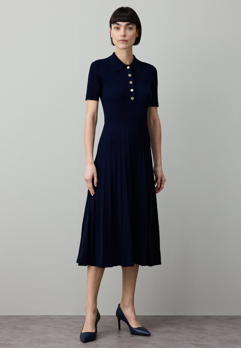 Temno modra obleka z rebrastim vzorcem, kratkimi rokavi, ovratnikom in gumbi. Razširjen, nabran krilo. Ujemajoči se čevlji z visokimi petami dopolnjujejo videz.