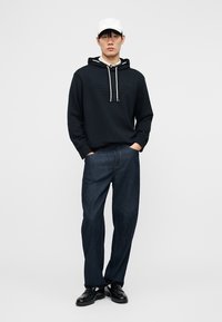 Homme portant une casquette blanche, un sweat à capuche noir avec des cordons, un jean bleu foncé et des chaussures en cuir noires, debout les mains dans les poches.