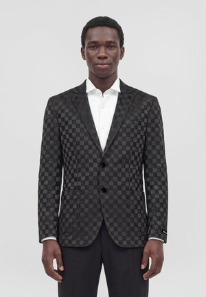 Un homme se tient face à l’objectif, vêtu d’une veste de costume sombre à carreaux, d’une chemise blanche et d’un pantalon noir, devant un fond uni et clair.