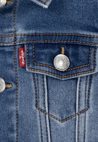 Denimjacka med ljusblått tyg, kopparstickningar och silverfärgade tryckknappar, med en bröstficka som har en detalj med röd etikett.