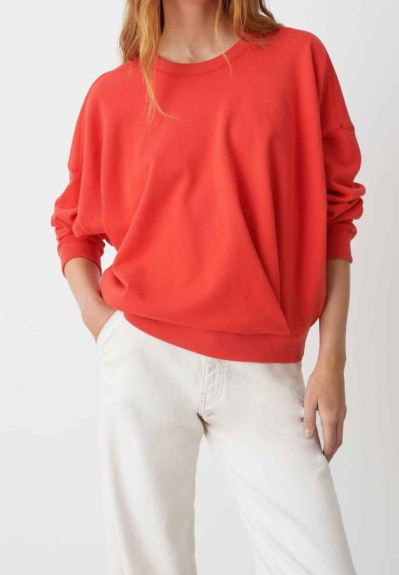 Sudadera roja de gran tamaño con puños y dobladillo acanalados, hecha de un material suave. Combinada con pantalones de corte relajado de color claro.