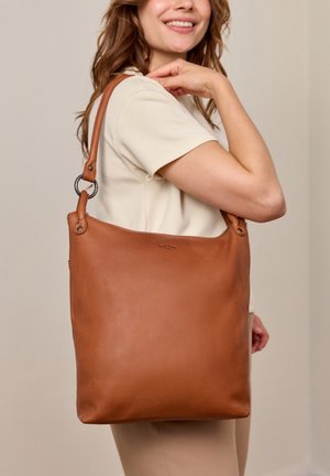 Woman smiles wearing a short-sleeve cream top, carrying a large smooth brown leather shoulder bag with metal rings on the straps.